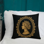 Decorative pillow with a gold portrait design on a black background, featuring a woman's profile and tiara, placed on a white surface with a teal wall in the background.